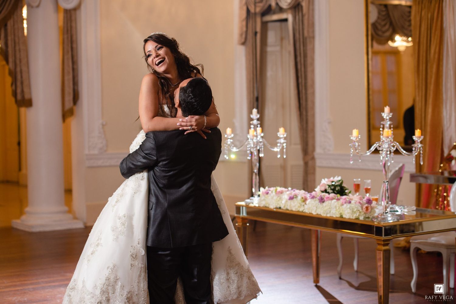 Bride and groom share a joyful embrace near a floral wedding cake table in an elegant ballroom.