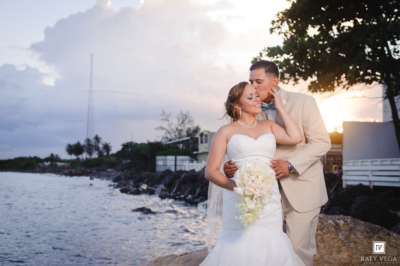 Bride and groom share a romantic sunset moment by the waterfront, bride in strapless gown holding white bouquet.