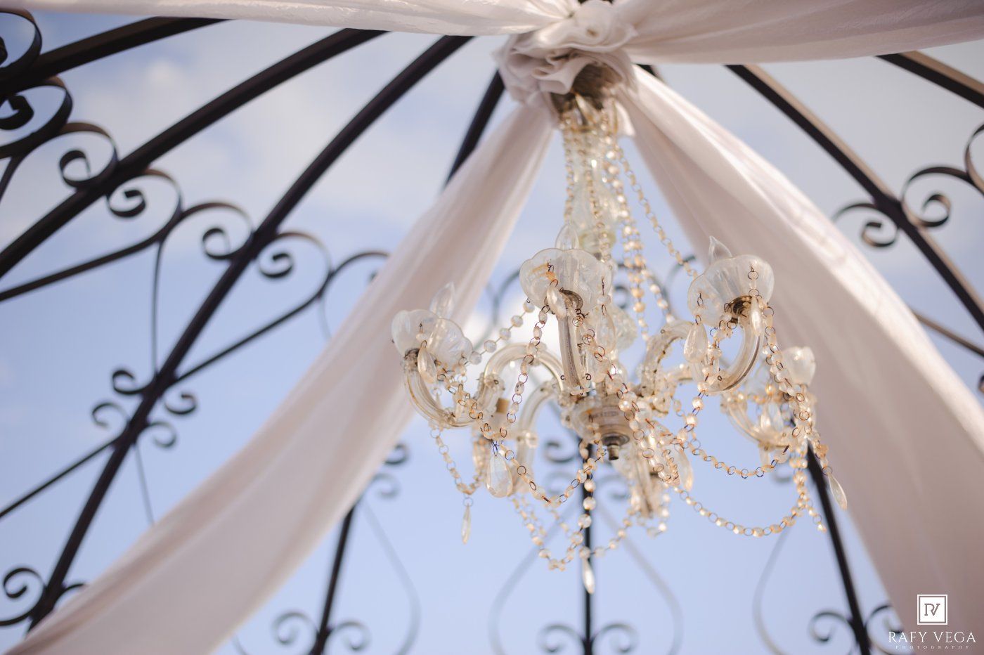 Crystal chandelier hanging from white draped fabric ceiling with ornate black ironwork gazebo frame against blue sky.