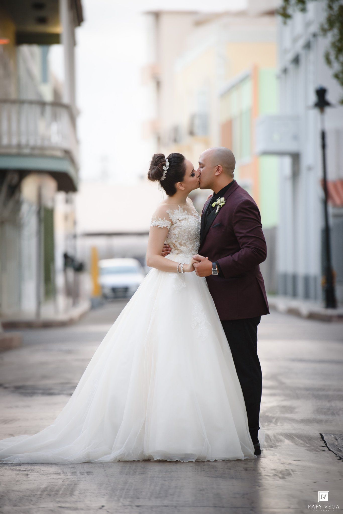 A bride and groom share a romantic kiss on a cobblestone street, she in a white ballgown, he in a burgundy suit.