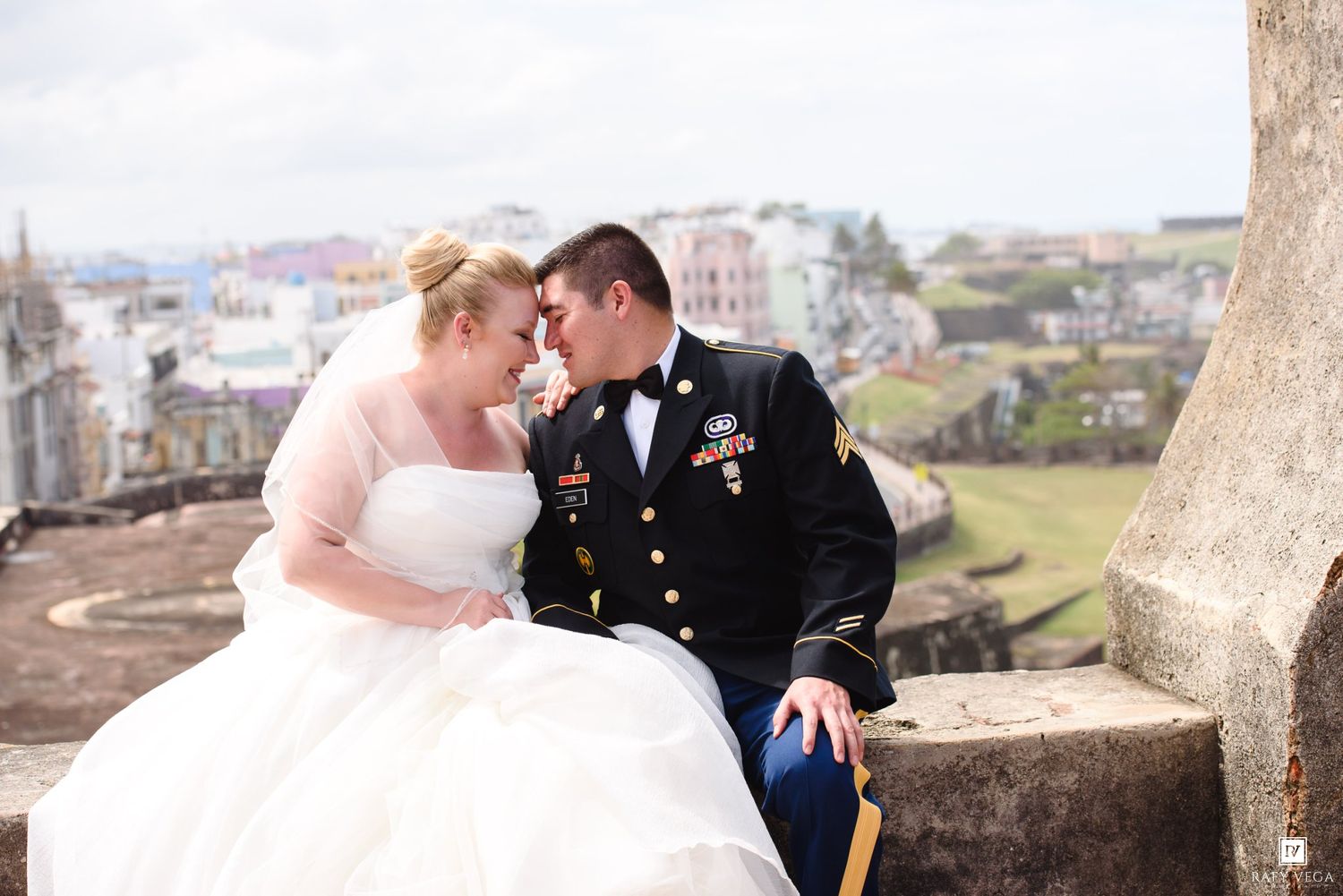 Military groom in dress uniform and bride in white gown share romantic moment seated on stone wall with colorful cityscape behind.