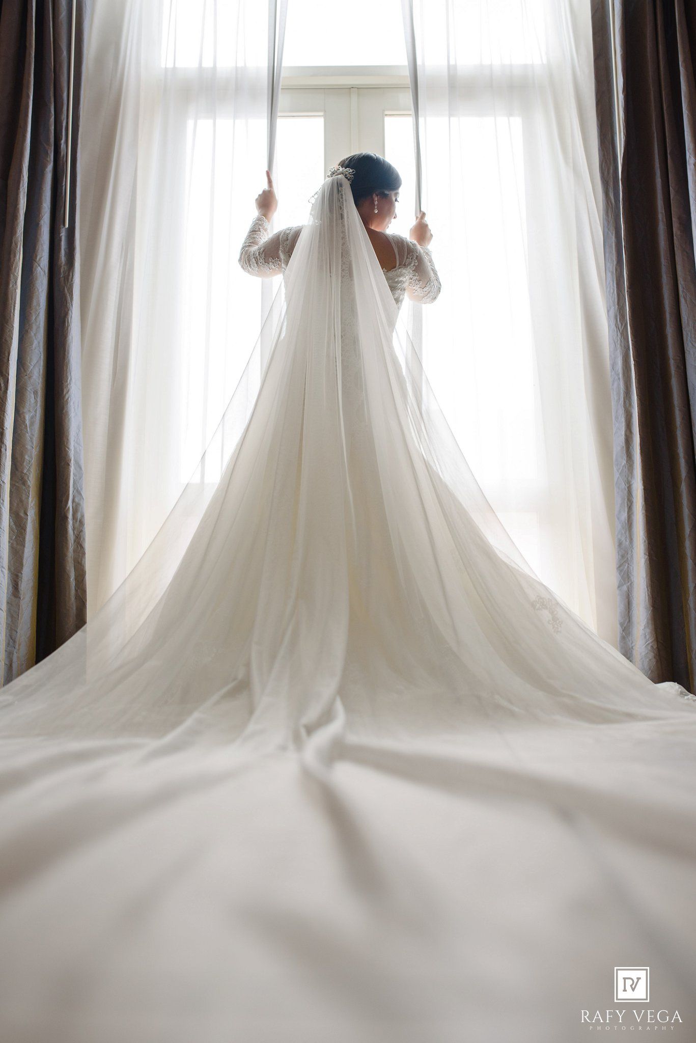 Bride in a flowing white wedding gown and long veil stands backlit by a bright window, holding curtains open.