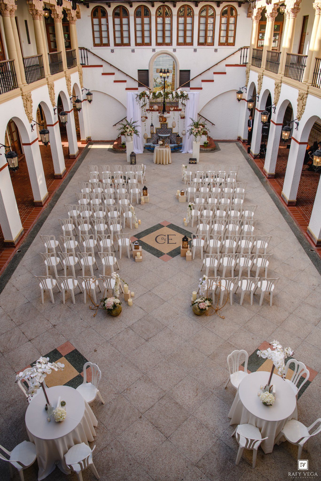 Aerial view of an elegant indoor wedding ceremony setup with white chairs, floral arrangements, and a decorated altar in a grand venue.