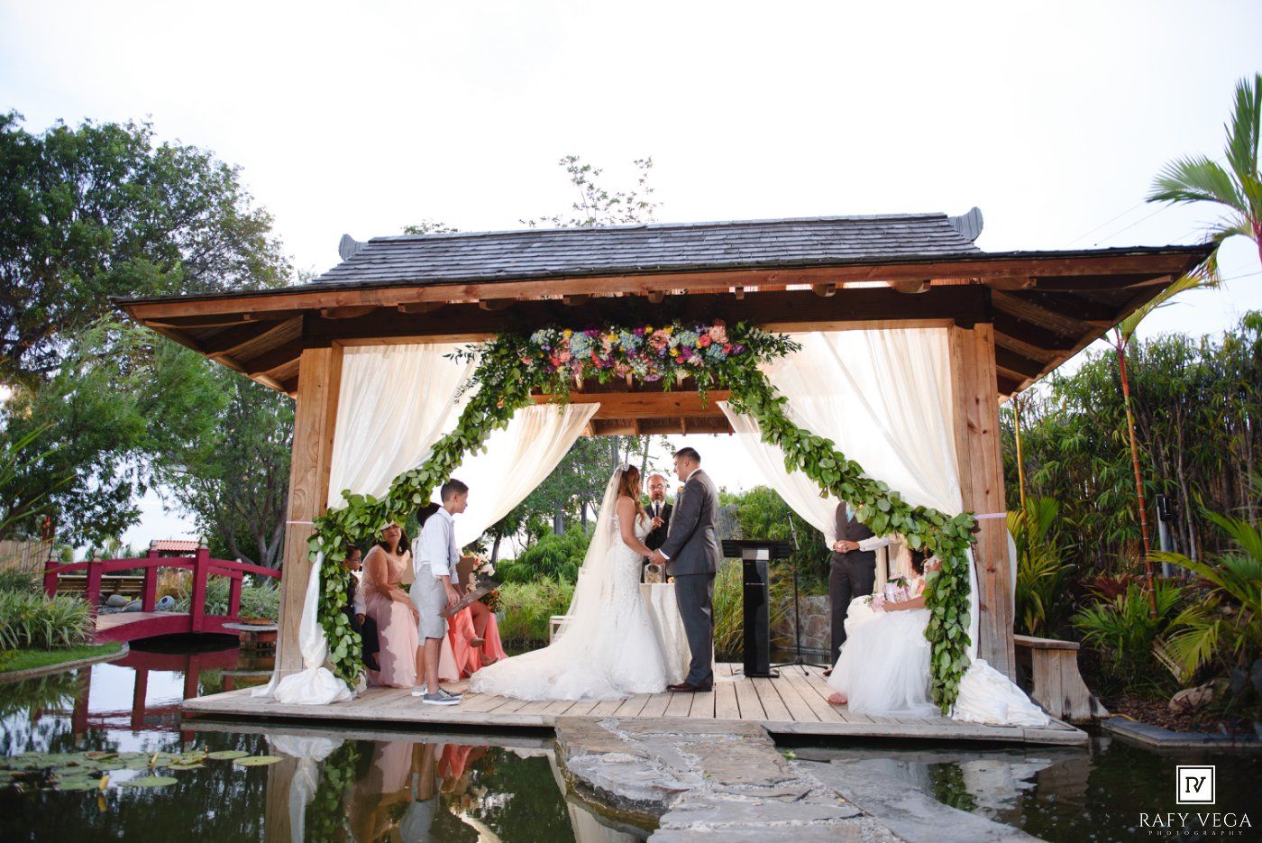 Outdoor wedding ceremony under a floral gazebo over a pond, with bride, groom, and wedding party on a wooden deck.