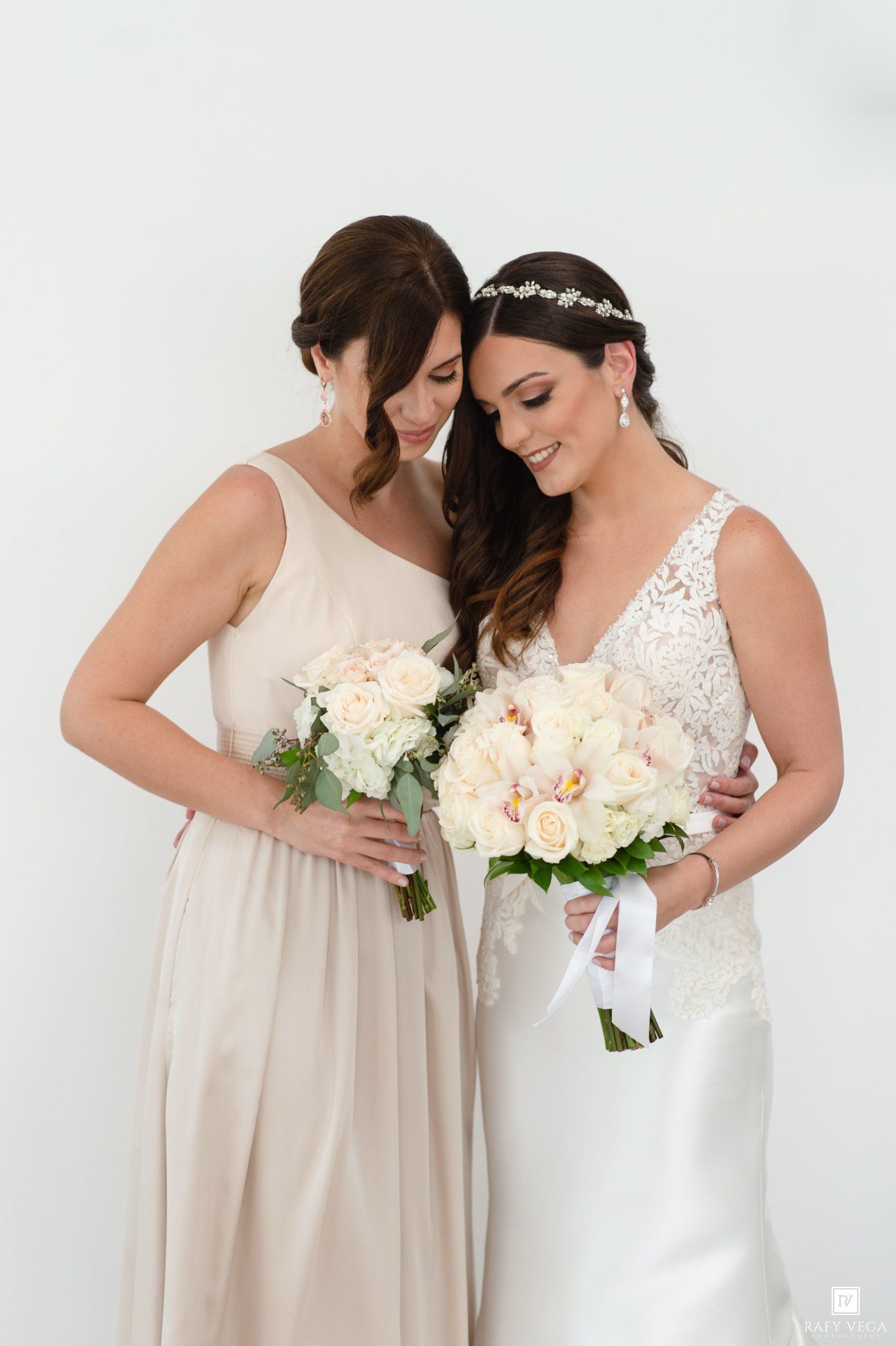 Bride in lace gown and bridesmaid in champagne dress holding white floral bouquets, sharing a tender moment.
