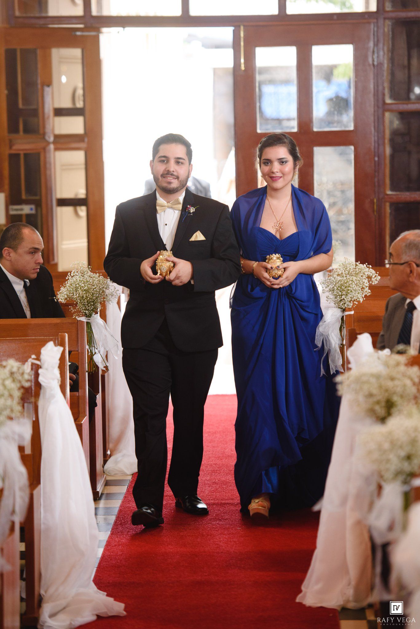 Couple walks down red carpet aisle in church wedding, man in tuxedo and woman in royal blue gown.