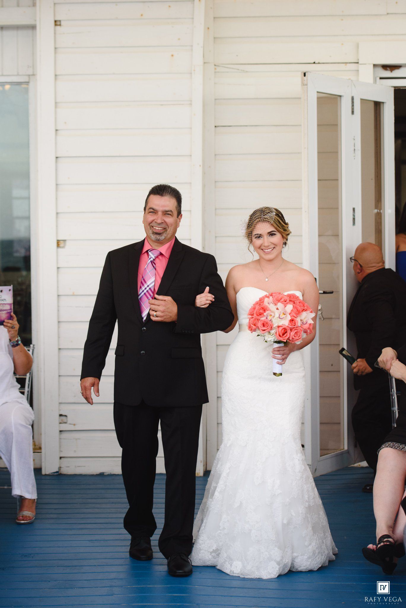 Bride in white mermaid gown holding coral bouquet walks with man in black suit on blue porch at outdoor wedding ceremony.