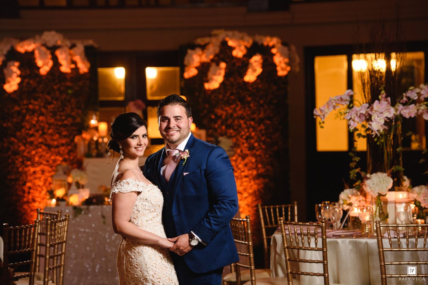 Elegant wedding couple posing at a romantic reception venue with warm amber lighting and floral arrangements.