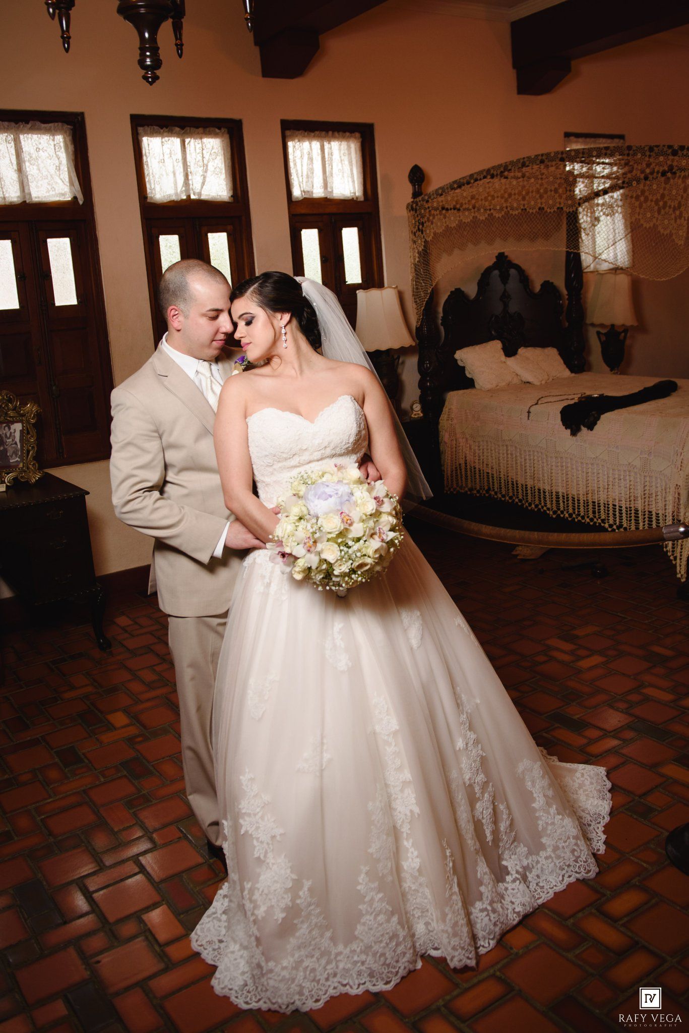 Bride in lace ball gown holding bouquet stands with groom in tan suit in elegant rustic room with ornate canopy bed.