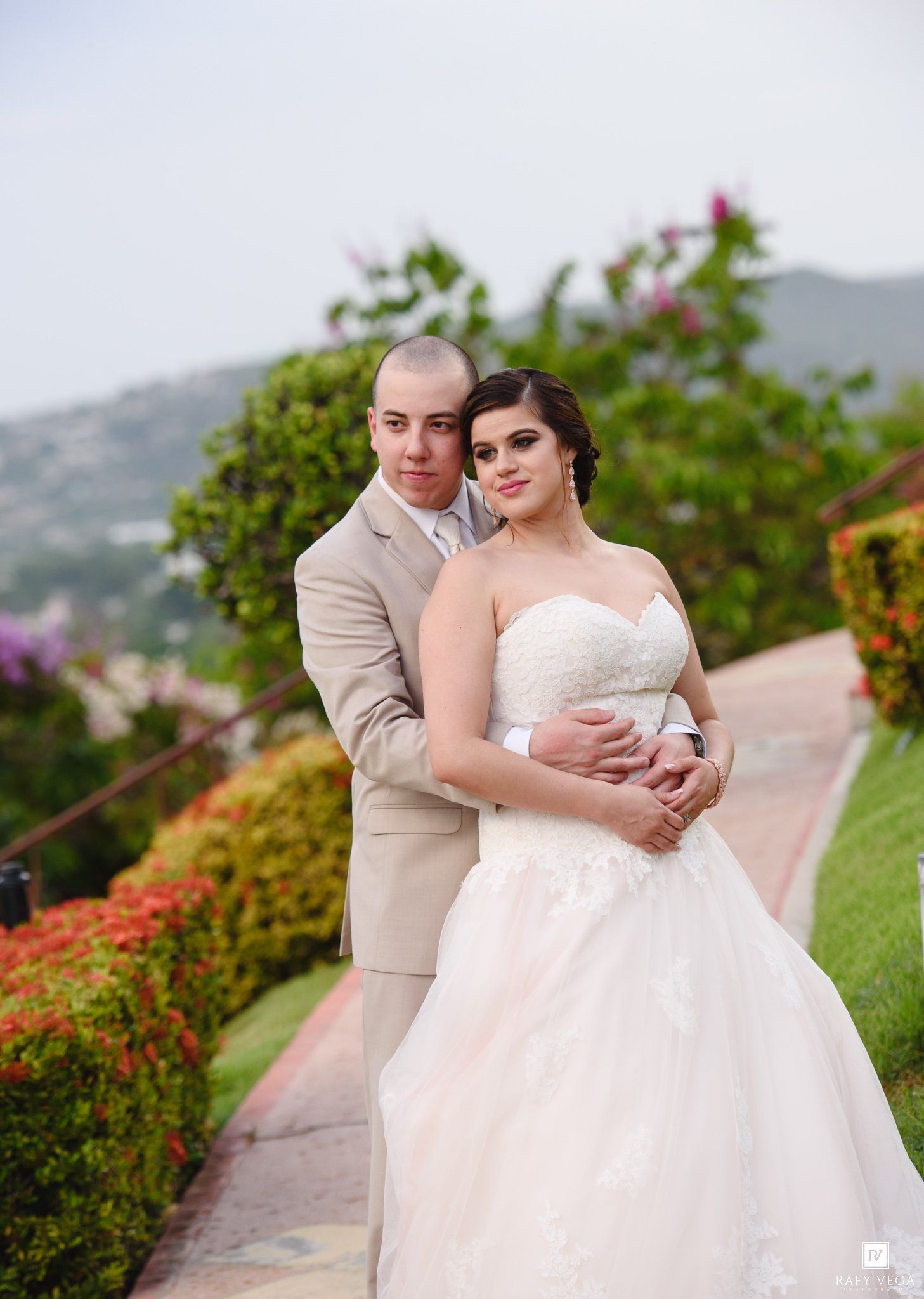 Bride in white strapless gown and groom in tan suit embrace outdoors at scenic garden wedding venue with mountain views.