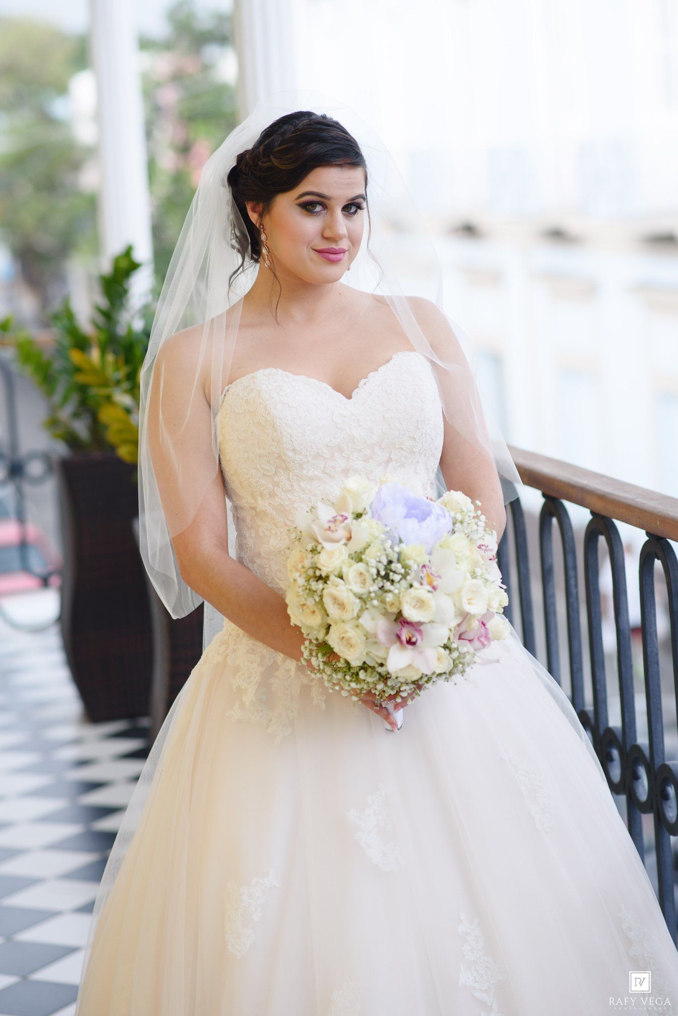 Bride in strapless lace ball gown with veil holding pastel floral bouquet on elegant venue balcony.