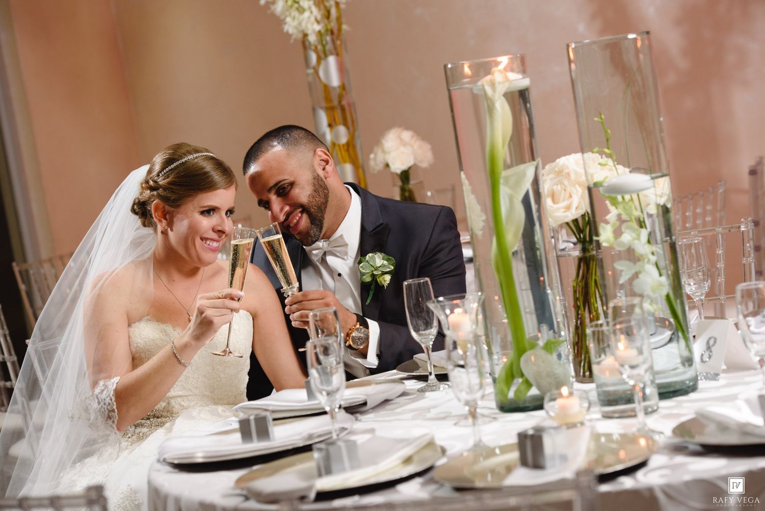 Smiling bride and groom toast with champagne glasses at their wedding reception table.