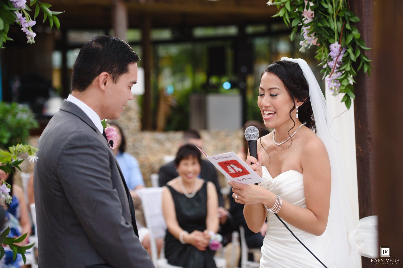 Bride reads wedding vows into microphone while groom listens during outdoor wedding ceremony.