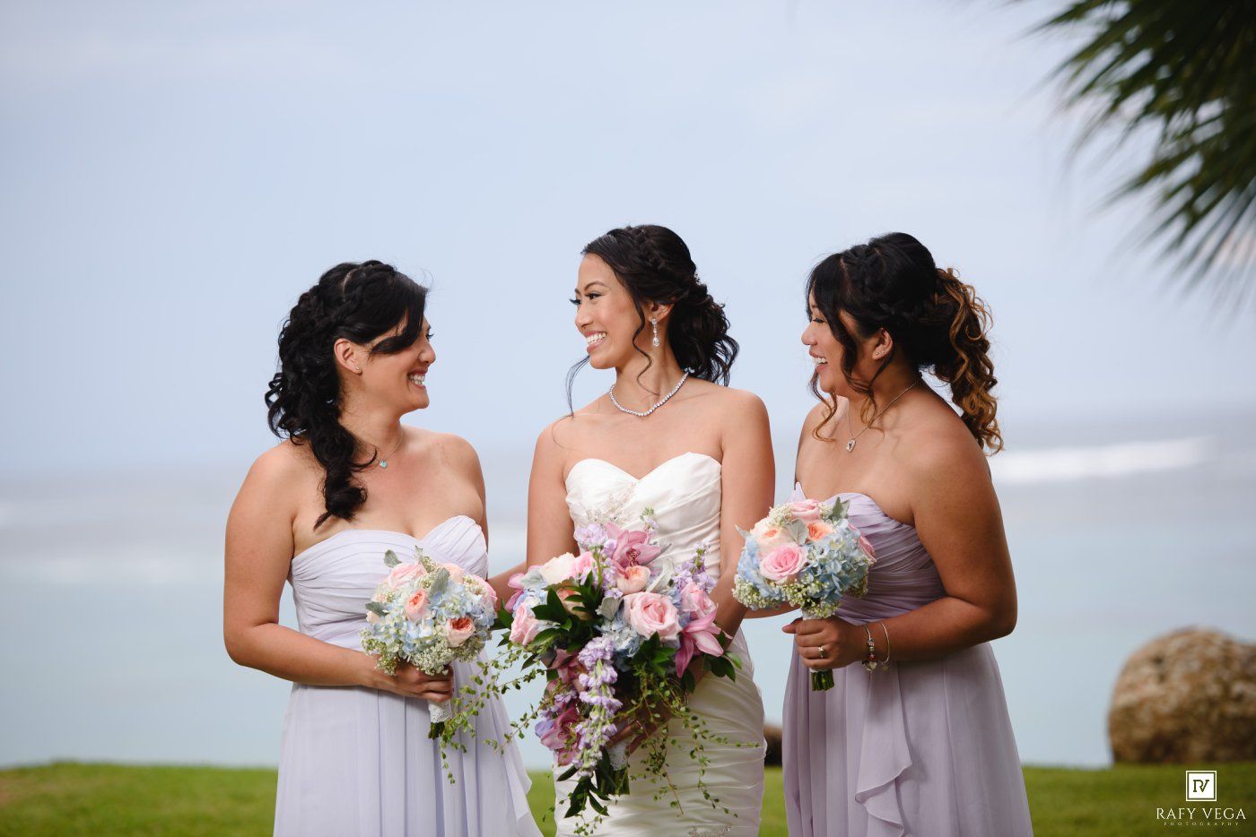 Bride in white gown holds bouquet flanked by two bridesmaids in lavender dresses at an outdoor beach wedding.