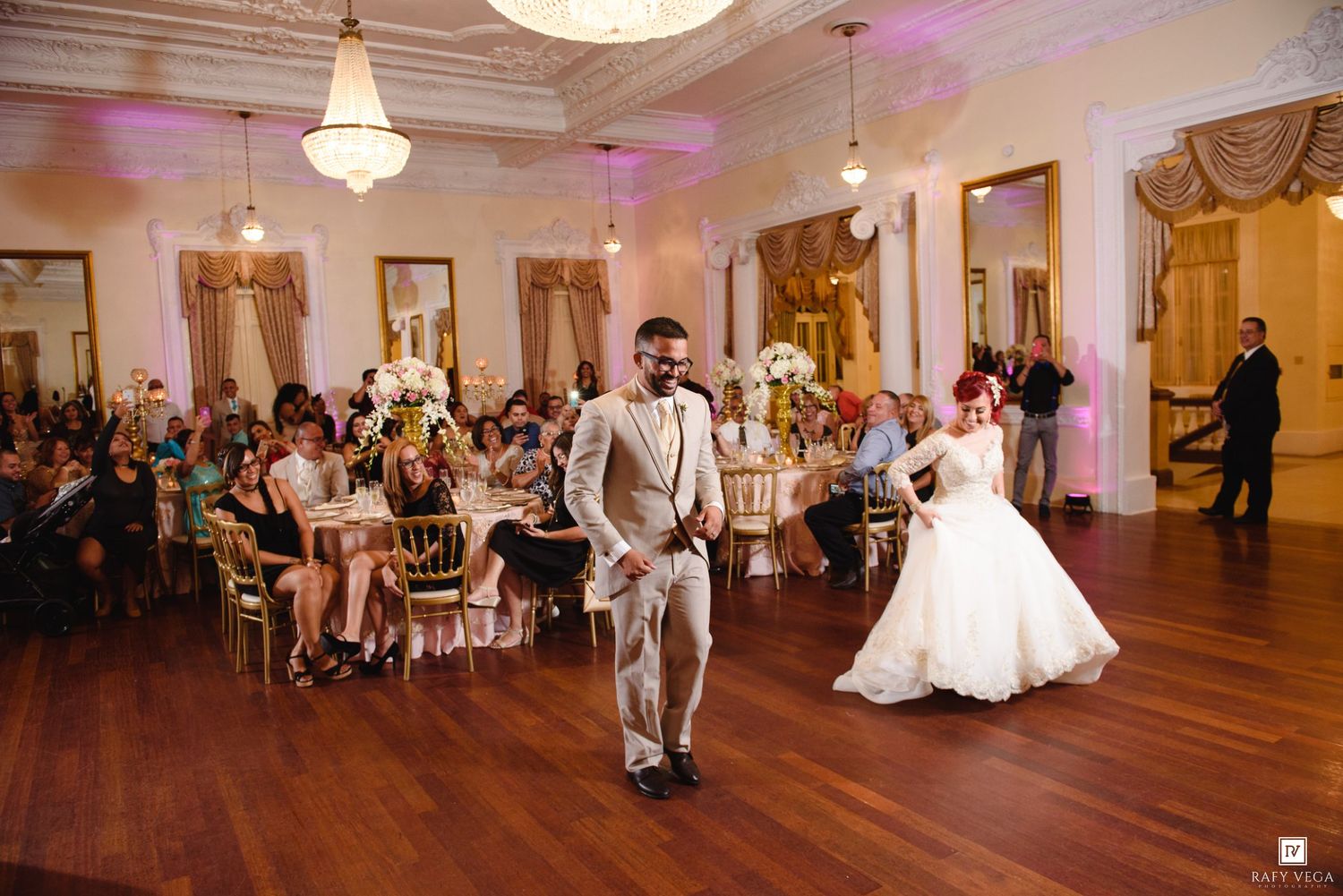 Bride and groom share first dance in elegant ballroom with guests seated at decorated tables during wedding reception.