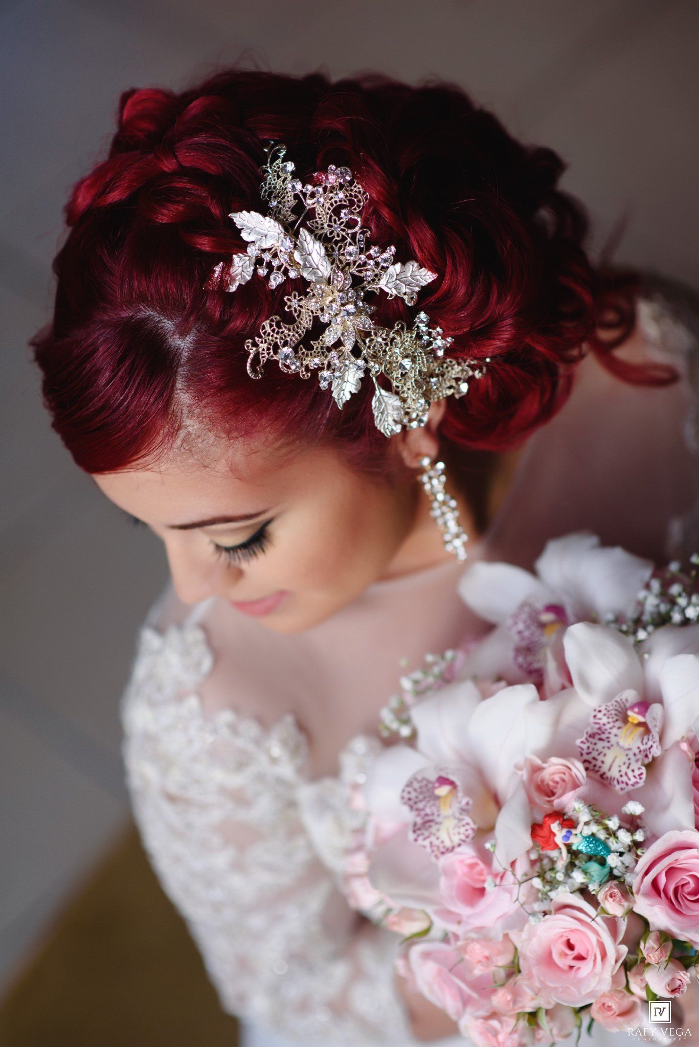 Bride with red updo hair adorned with crystal hair piece holding pink floral bouquet in white lace wedding dress.