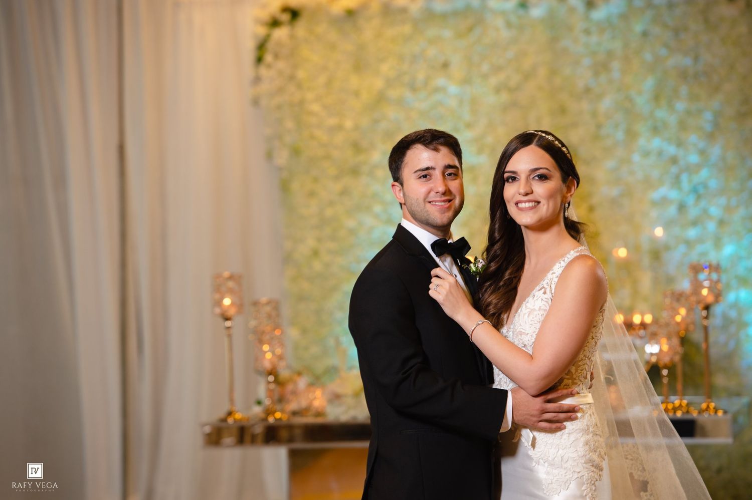Bride and groom posing elegantly in front of a floral wall backdrop at their wedding reception.
