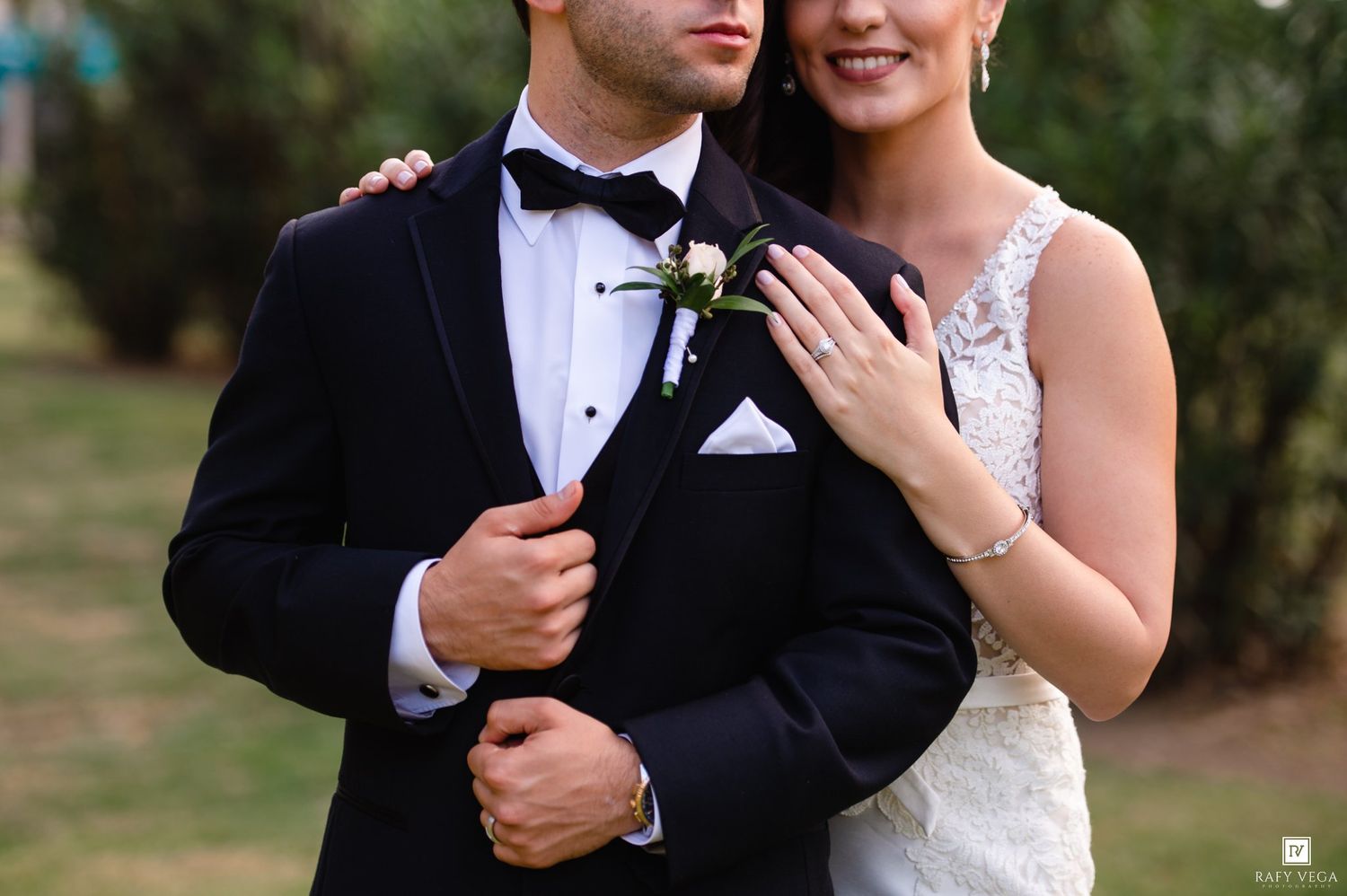 Groom in black tuxedo and bride in beaded gown embracing outdoors, showcasing her diamond engagement ring.