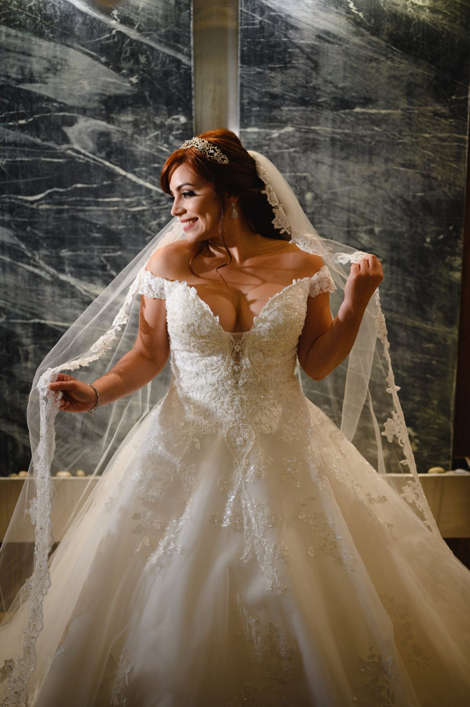 Bride in off-shoulder lace ball gown with veil and tiara, smiling elegantly near a marble wall.