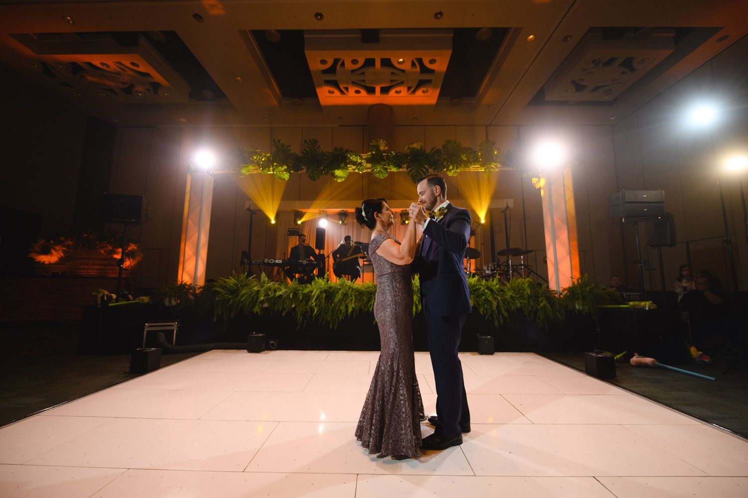 Couple sharing first dance on white dance floor at elegant wedding reception with warm amber lighting.