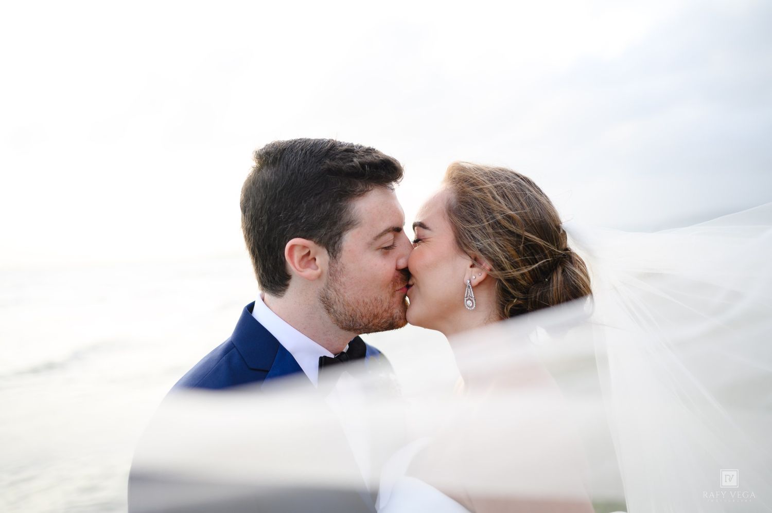 A bride and groom share a romantic kiss outdoors, with the wedding veil flowing around them against a bright sky.