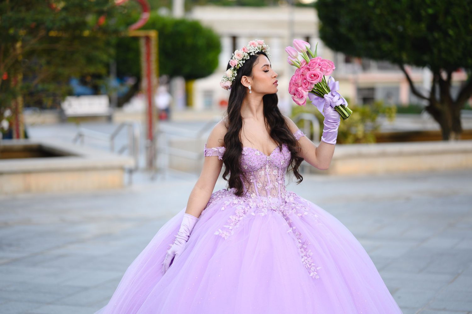Young woman in lavender quinceañera ball gown with floral crown holds pink bouquet outdoors.