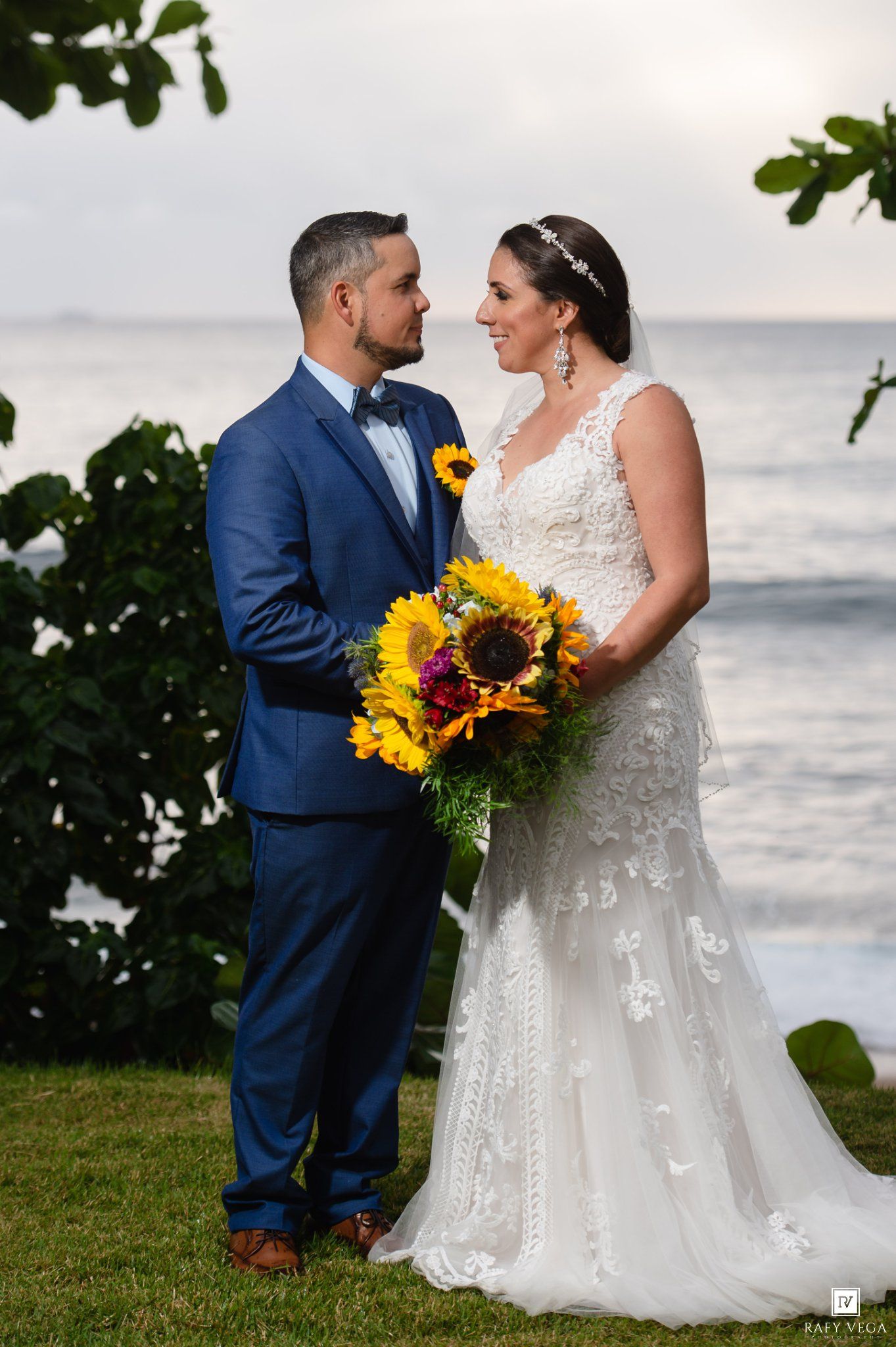Bride and groom share a romantic moment by the ocean, she holds sunflower bouquet in lace gown, he wears blue suit.