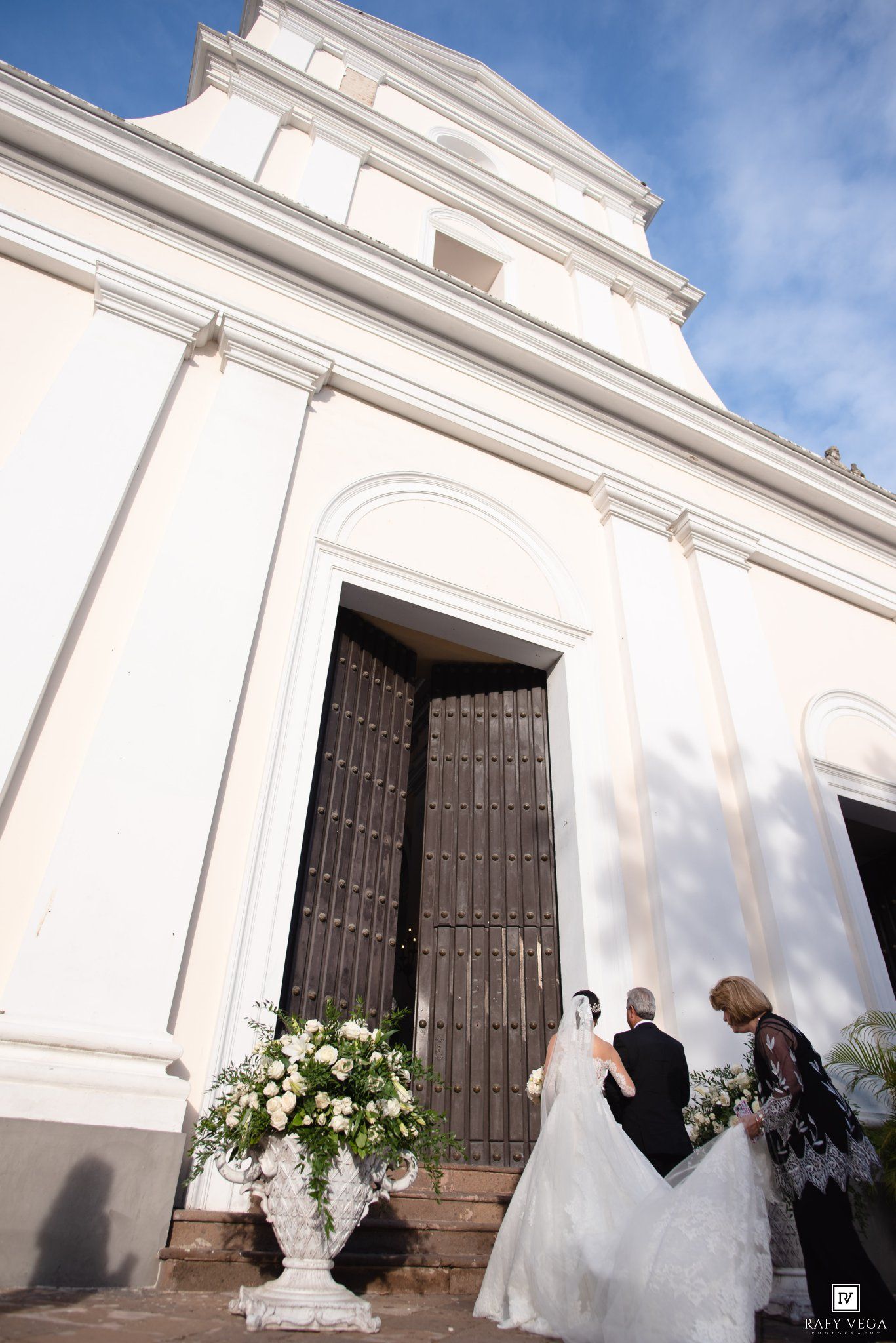 Bride in white gown arrives at elegant white church entrance with floral arrangement for wedding ceremony.