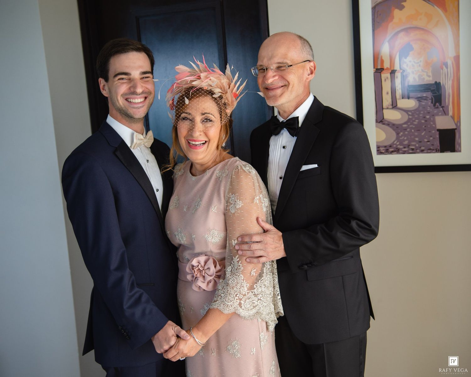 Family of three posing together at a wedding, woman in pink lace dress with floral hat, men in tuxedos.