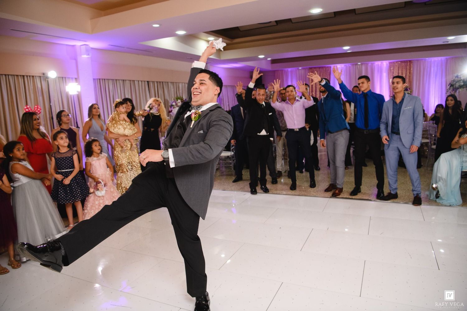 Groom dramatically throws bouquet to cheering wedding guests on white dance floor at elegant reception.