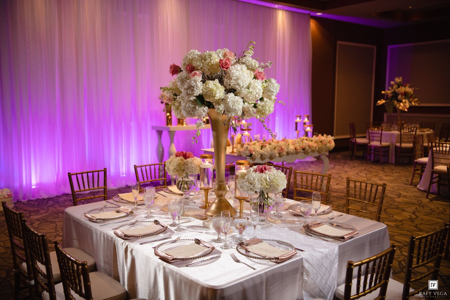 Elegant wedding reception with tall floral centerpieces, chiavari chairs, white linens, and purple ambient lighting.