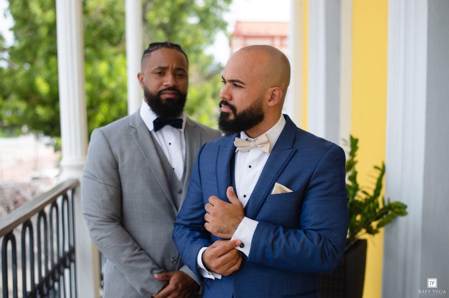 Two well-dressed grooms in blue and gray suits pose on a porch during their wedding ceremony.