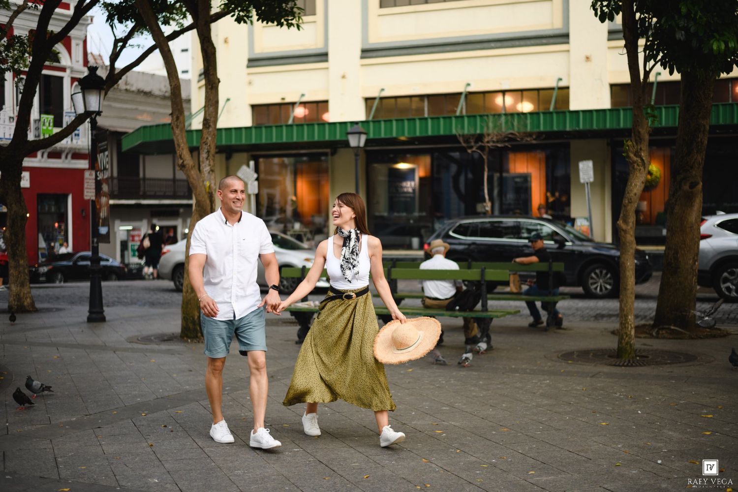Couple walking hand in hand through a charming urban plaza, woman in olive skirt holding a hat.
