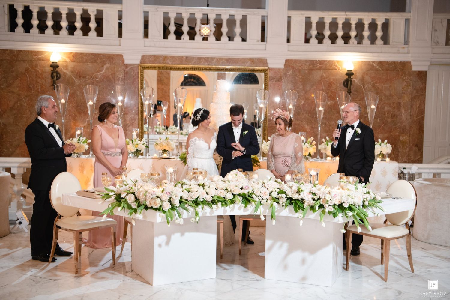 Elegant wedding reception with bride and groom at floral-adorned sweetheart table surrounded by family in grand marble venue.