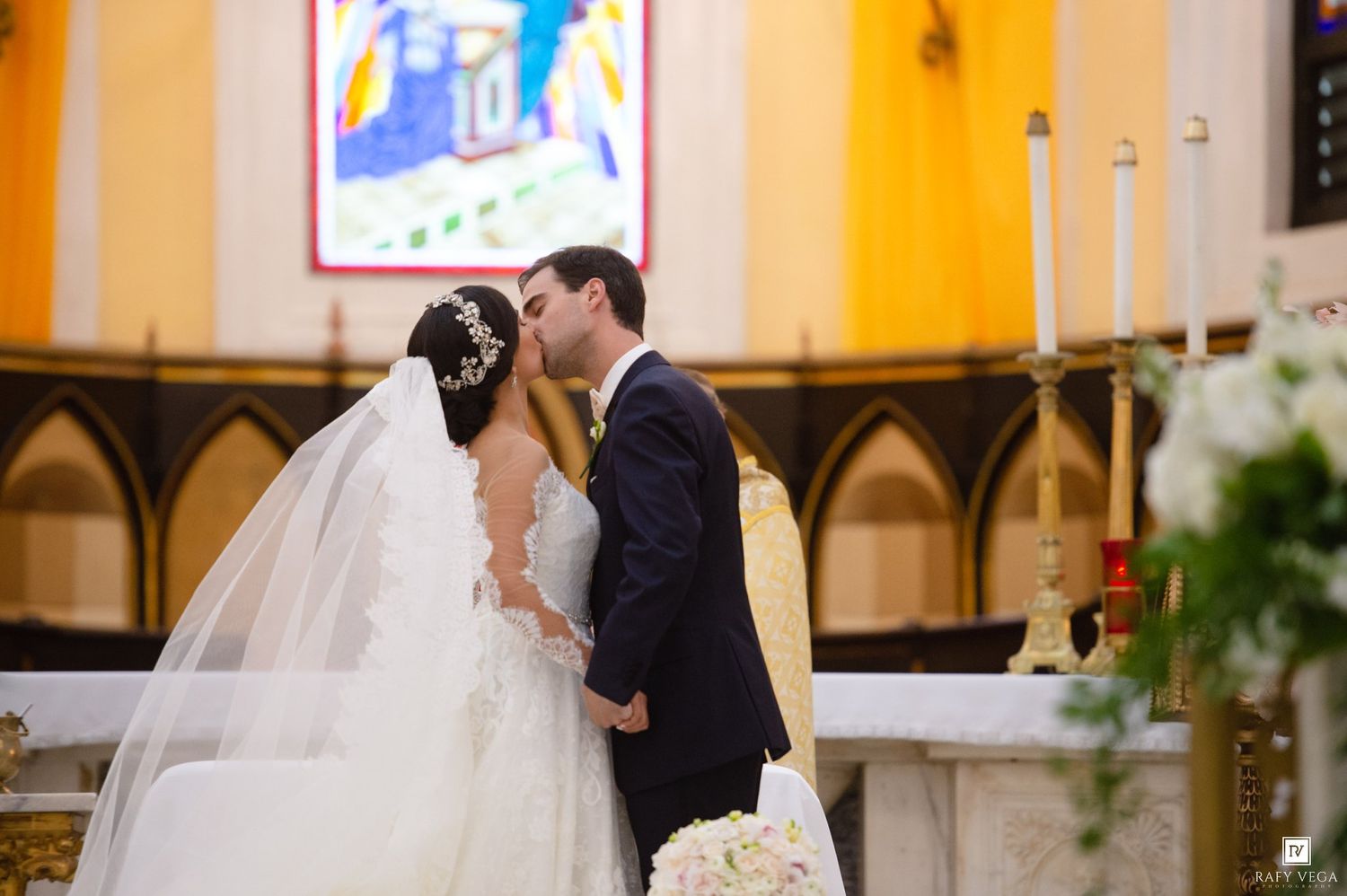 Bride and groom share first kiss at church altar during elegant wedding ceremony with stained glass windows.