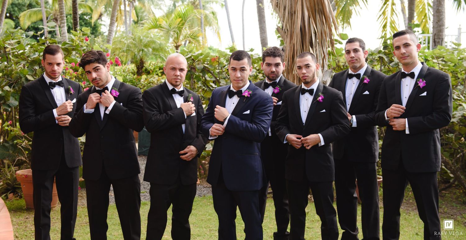 Groomsmen in black tuxedos with pink boutonnieres pose outdoors at a tropical wedding venue.