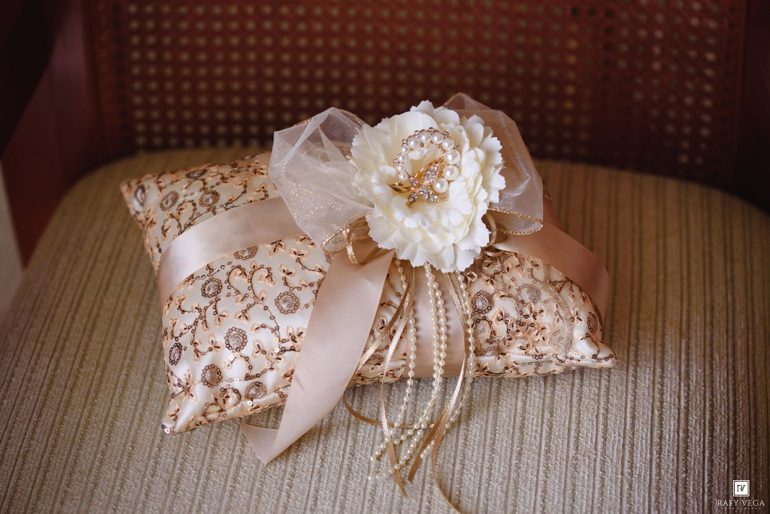 Elegant wedding ring pillow with gold brocade fabric, blush satin ribbon, white peony flower, and pearl accents on a chair.