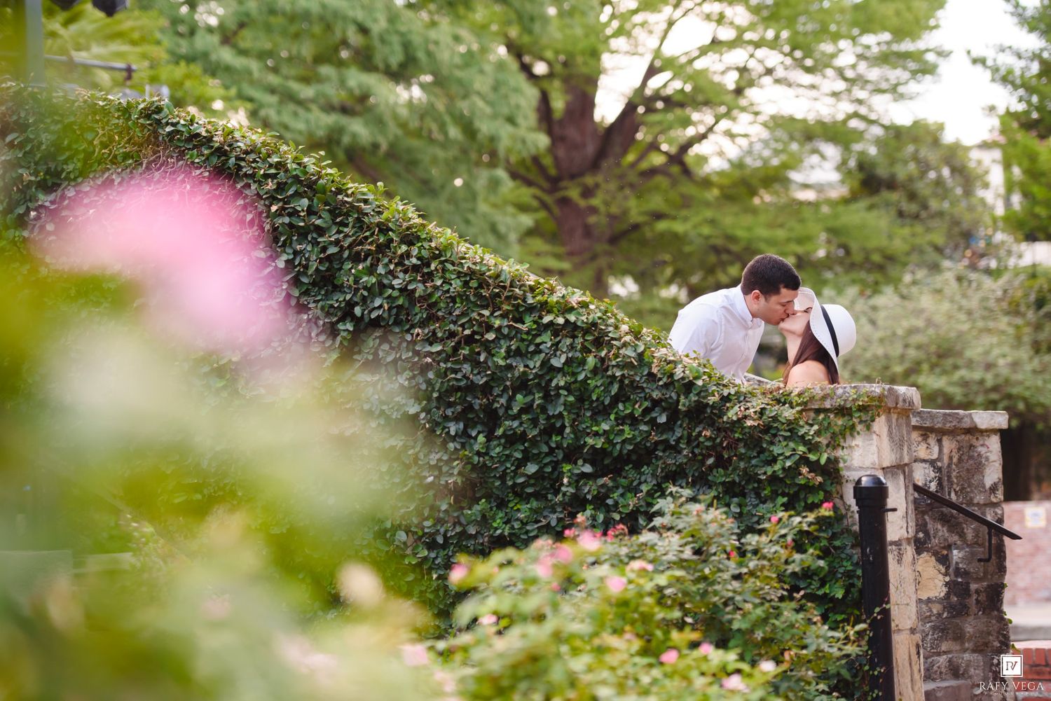 Couple sharing a romantic kiss over an ivy-covered stone wall surrounded by lush gardens and pink flowers.
