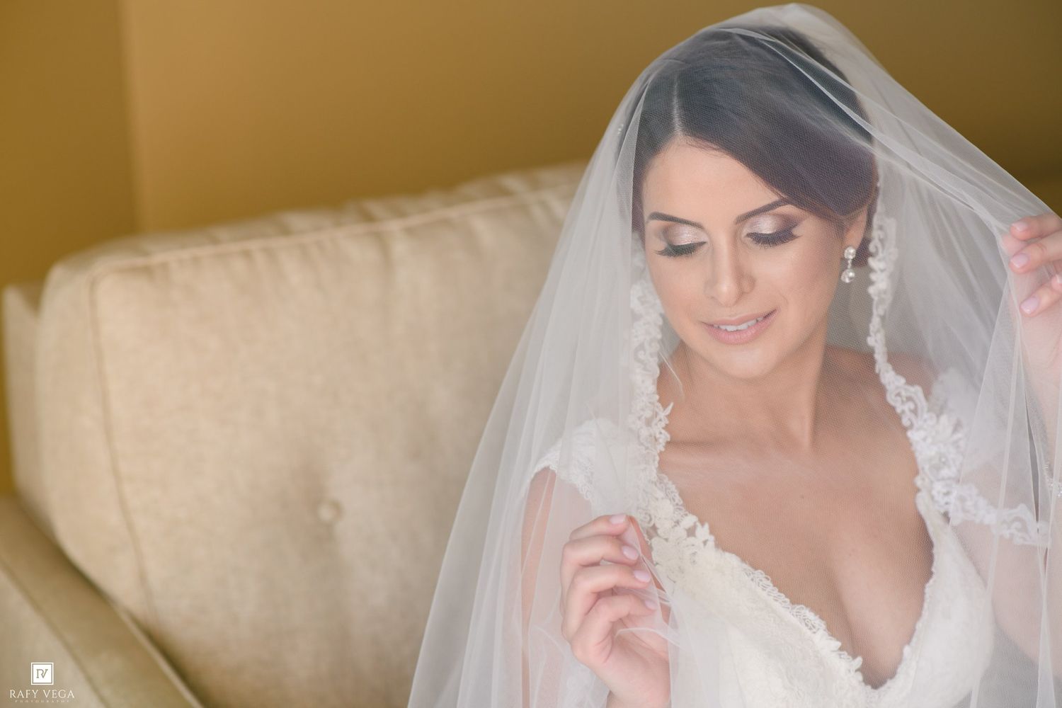 Bride in white lace wedding dress and veil smiling down, wearing elegant earrings, seated on a beige sofa.