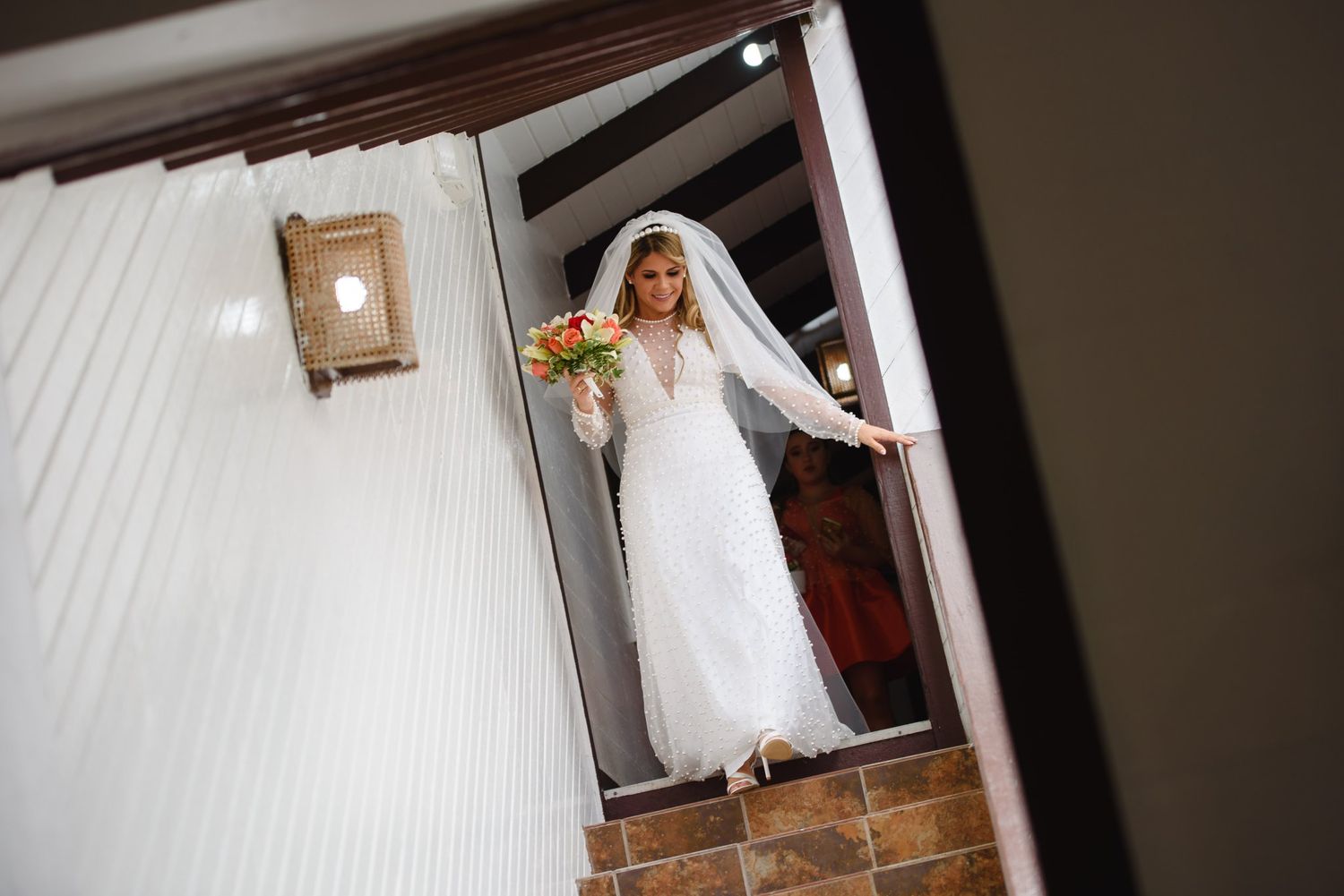 Elegant bride in white gown and veil descends staircase holding colorful bouquet at wedding venue.