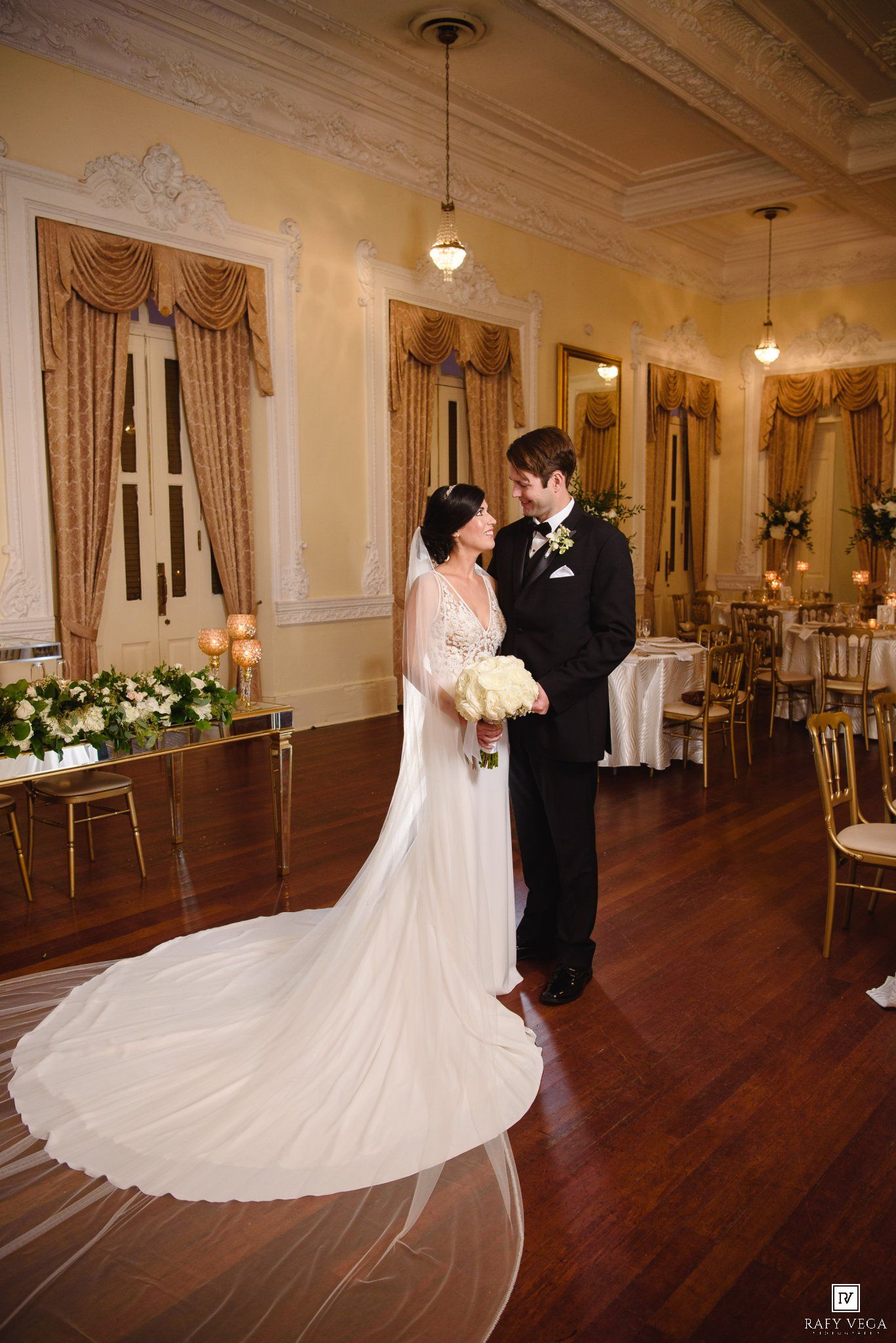 Bride and groom share a romantic moment in an elegant ballroom with gold decor, hardwood floors, and a flowing white wedding gown.