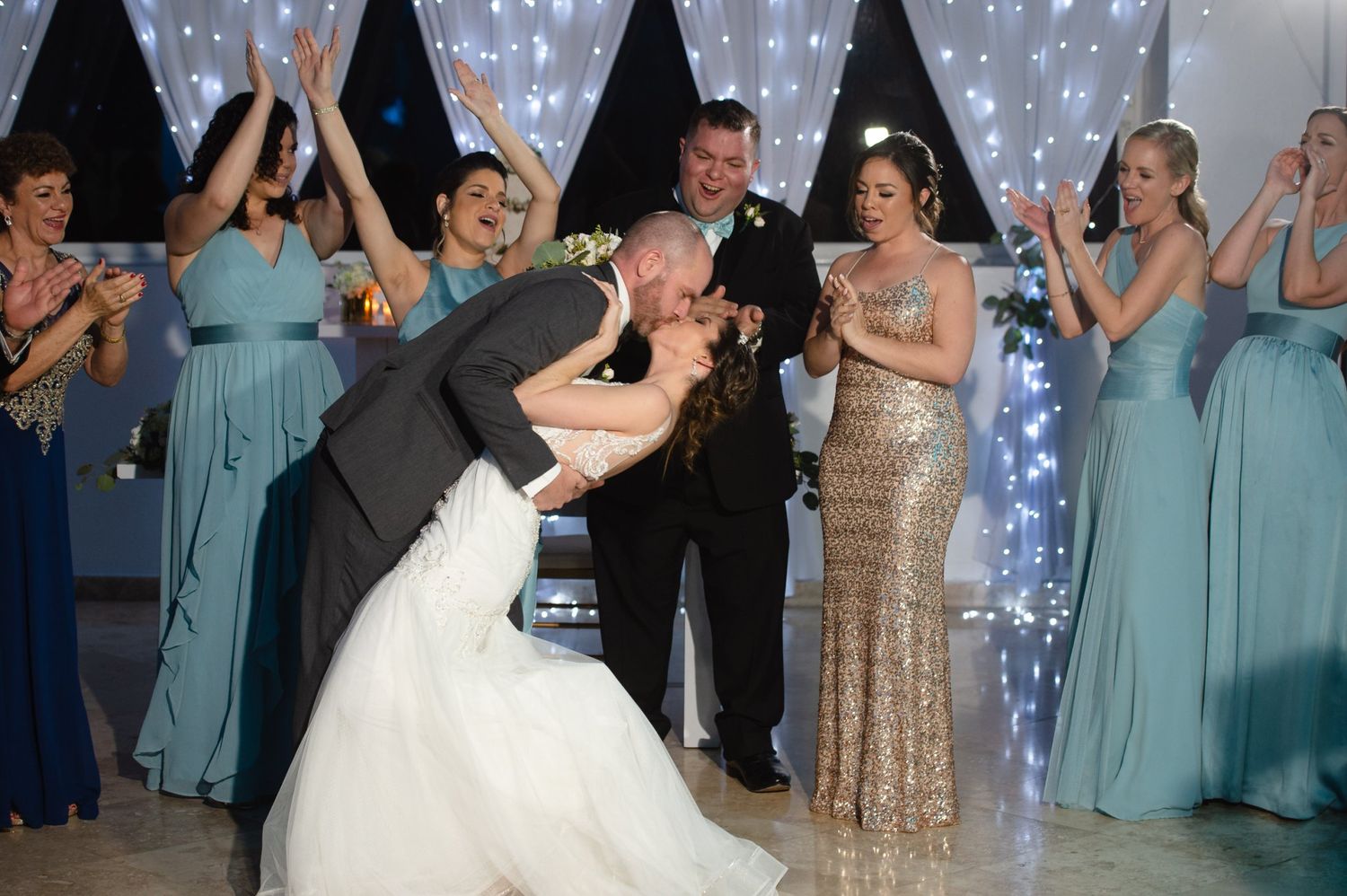 Groom dips bride for a kiss on the wedding dance floor surrounded by cheering bridesmaids in teal gowns.
