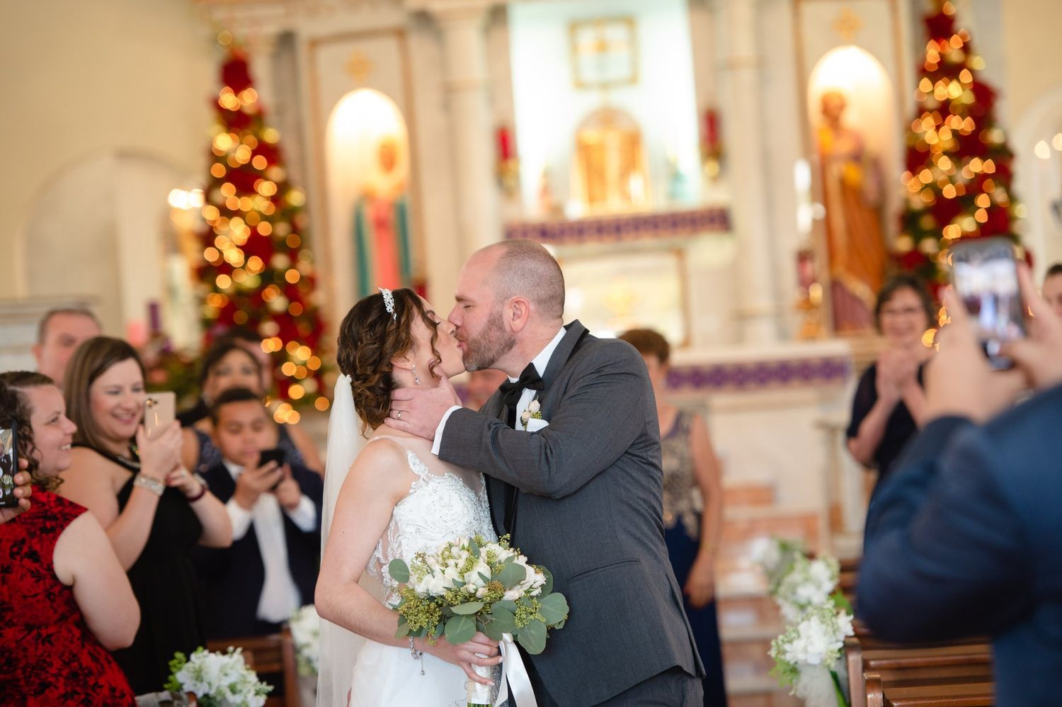 Bride and groom share first kiss at church altar with Christmas trees and guests celebrating in background.