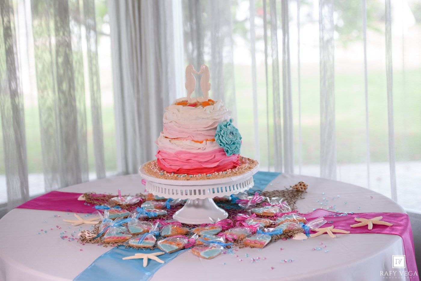 Pink ombre wedding cake on white stand surrounded by starfish and colorful beach-themed decorations on a round table.