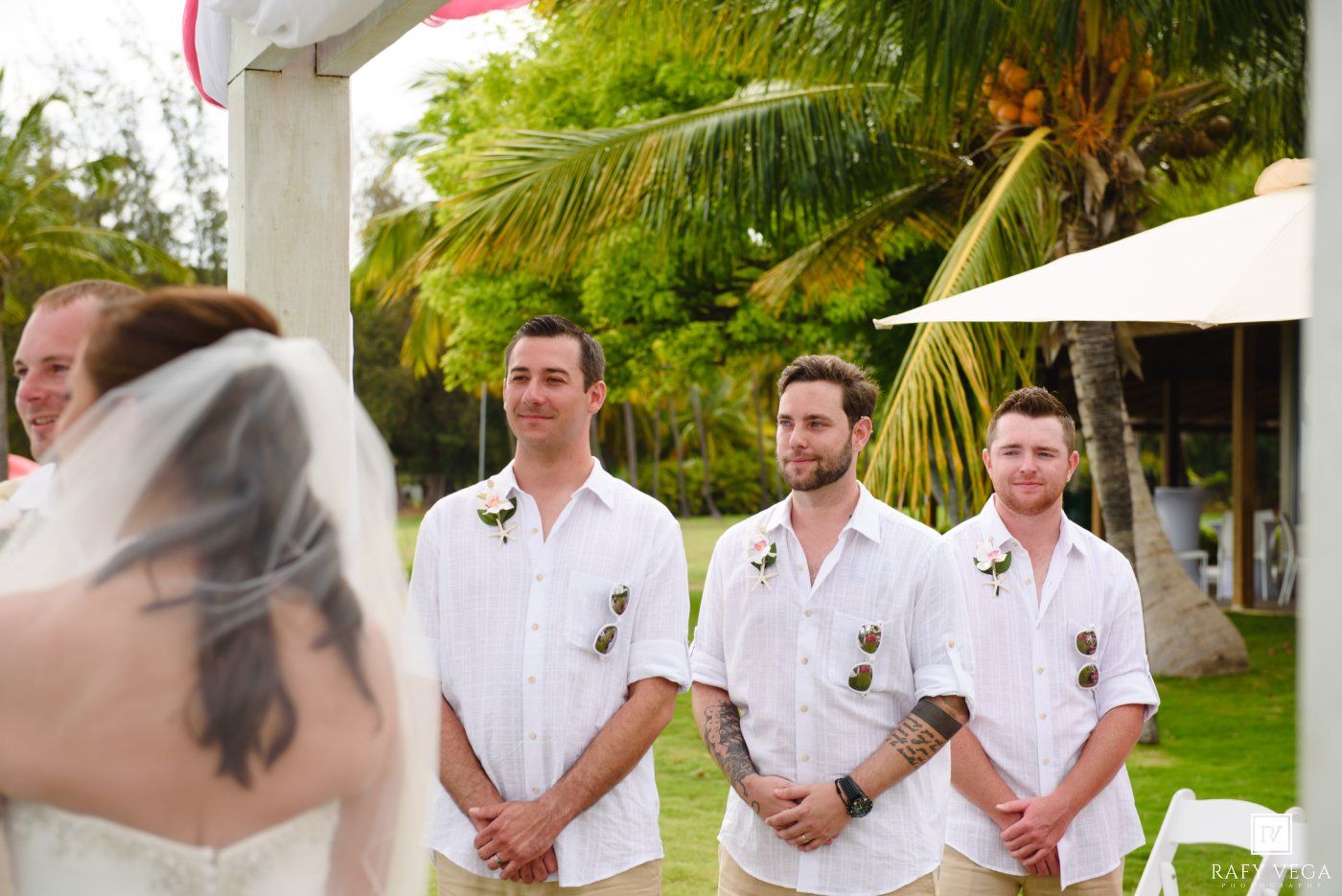 Groomsmen in white shirts watch the bride approach during an outdoor tropical wedding ceremony with palm trees.