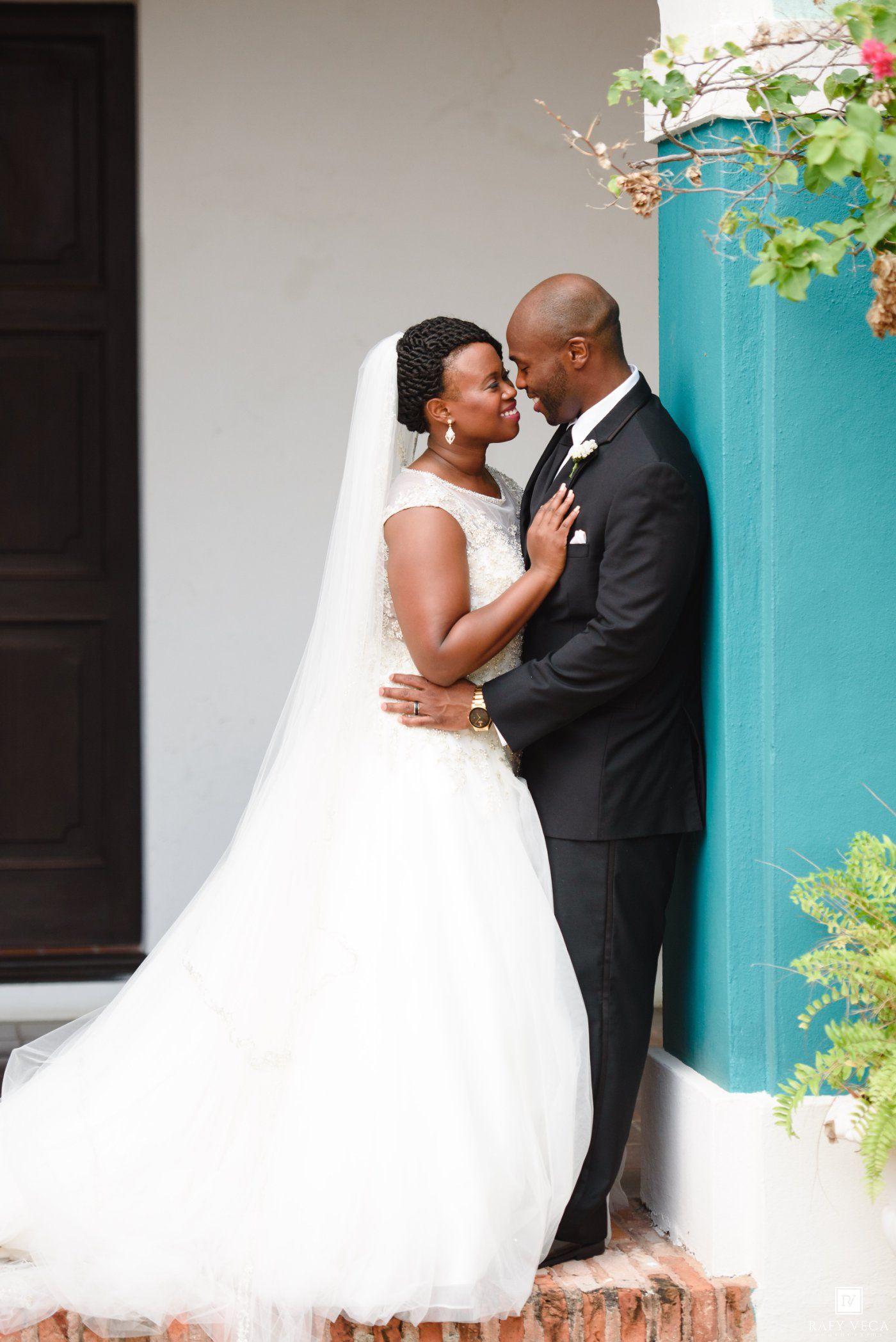 Bride and groom share a romantic moment leaning against a teal wall, bride in white ballgown with veil.