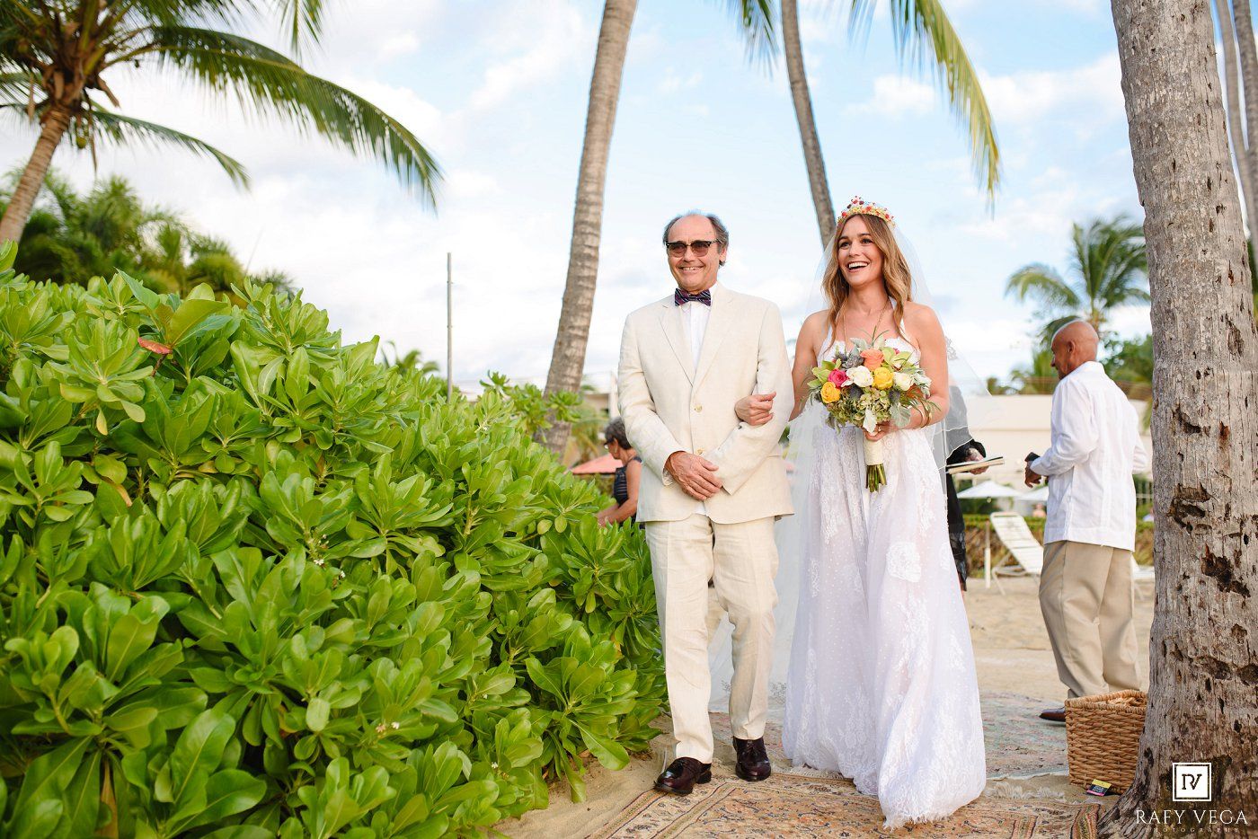 Bride in white gown walks with escort along tropical beach path lined with palm trees and lush greenery at outdoor wedding.