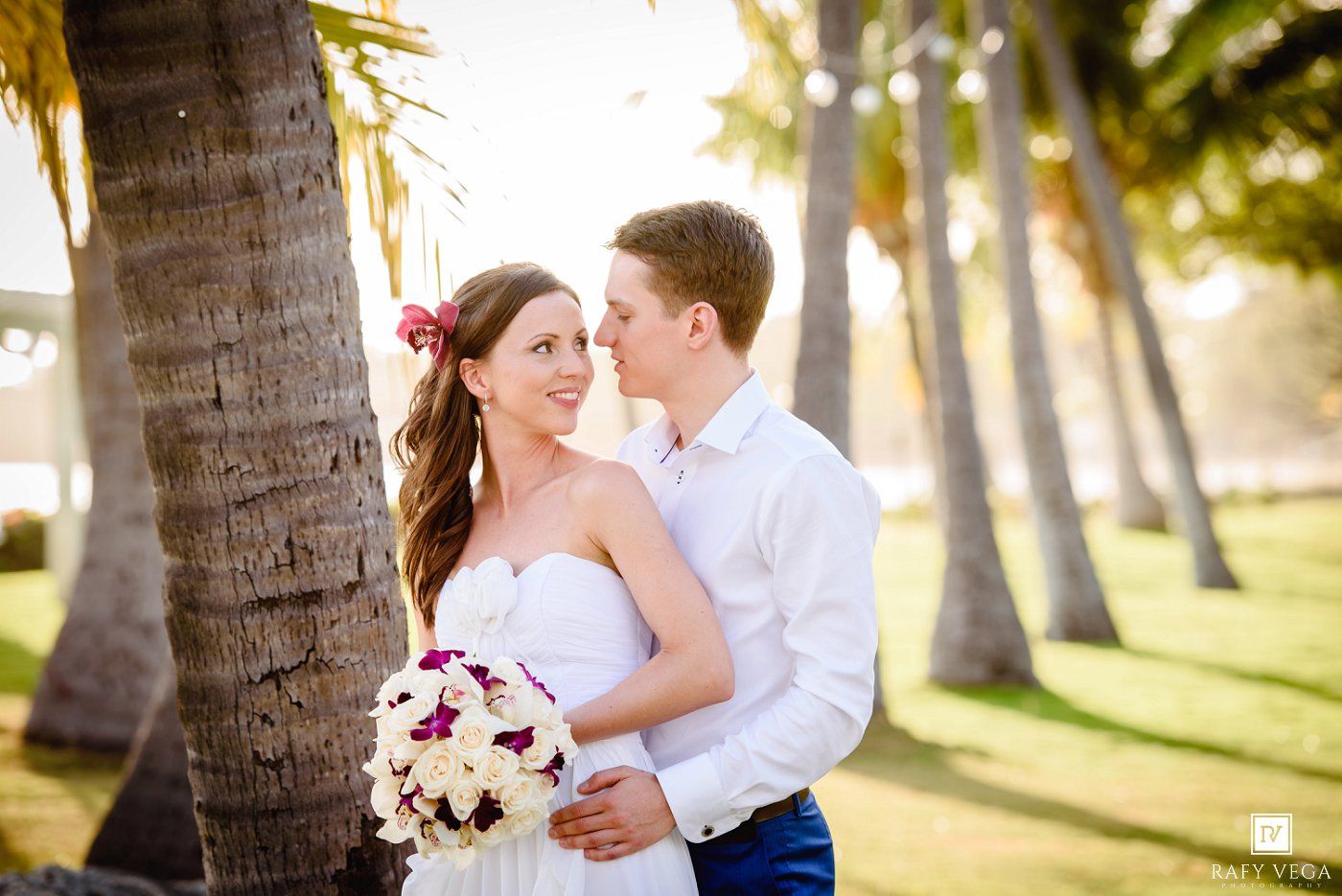 A bride and groom share a romantic moment among palm trees at a tropical beach wedding.