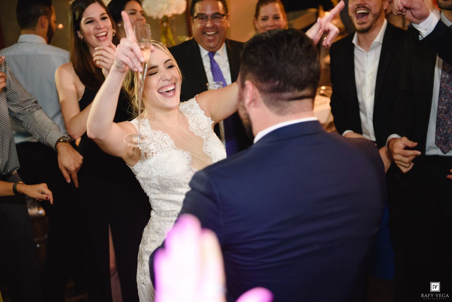 Bride in lace wedding gown laughing and dancing joyfully with groom and guests at lively wedding reception.