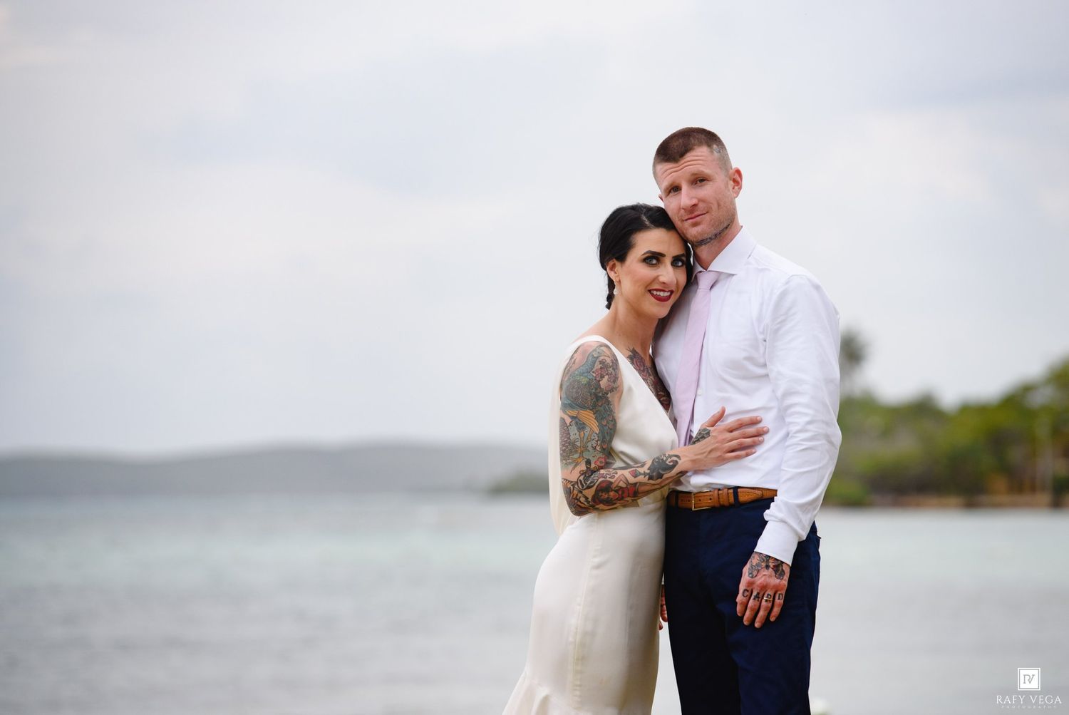 Couple in wedding attire embracing on a tropical beach, bride with sleeve tattoos in white gown, groom in white shirt.